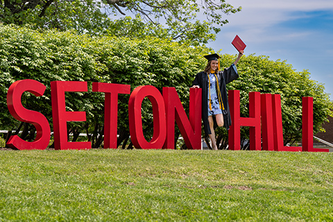 A graduate standing next to large letters that say Seton Hill