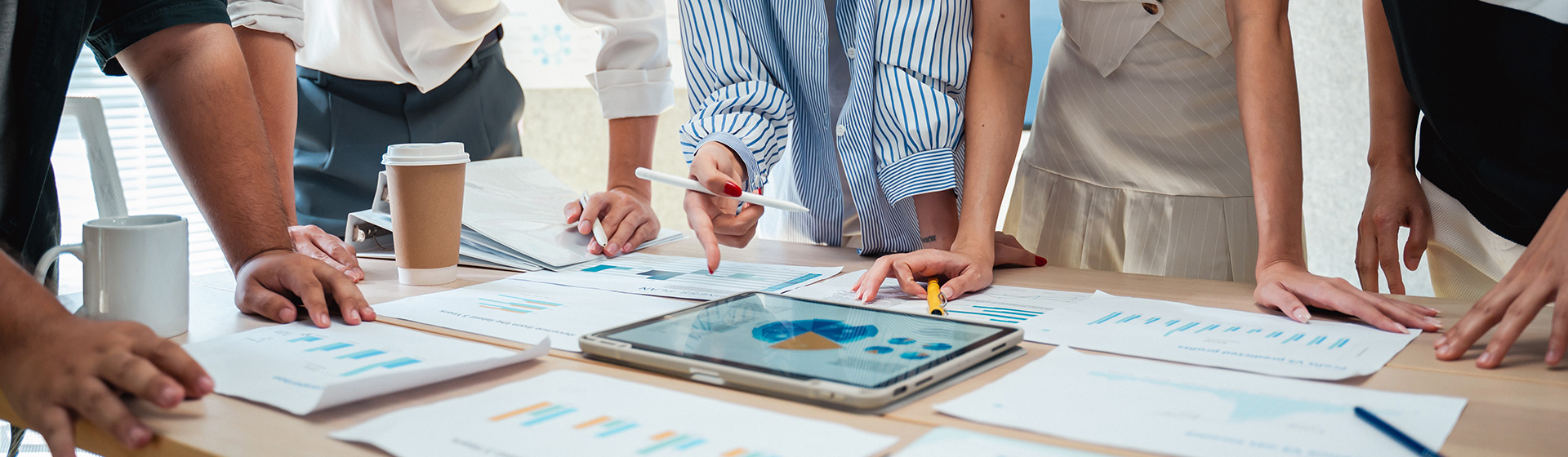 A team of people lean over a table with an digital tablet in the center surrounded by charts and graphs printed on paper.